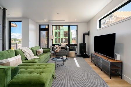 A modern living room with a green sectional sofa, a flat-screen TV on a stand, a coffee table, and large windows letting in natural light. A door leads to a balcony with a view of a construction site outside.