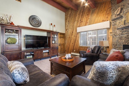 A cozy living room features a television in a wooden entertainment center, a coffee table, and comfortable seating with plush pillows. The space has wood-paneled walls and a stone fireplace.