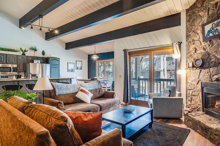 A modern living room with exposed beams, a stone fireplace, brown sofas, a coffee table, and a view of a dining area and a kitchen in the background.