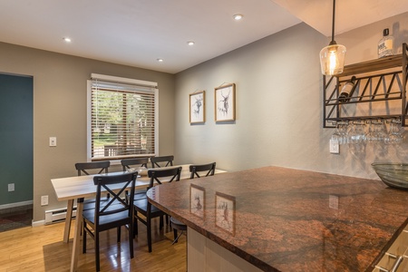 A dining area with a table and six chairs beside a window, adjacent to a kitchen counter with a hanging wine glass rack. Two framed pictures are on the wall.