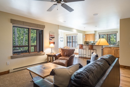 A cozy living room with a leather sofa, armchair, wooden coffee table, and a ceiling fan. The room has large windows, a view of trees, and an open kitchen with wooden cabinets and granite countertops.