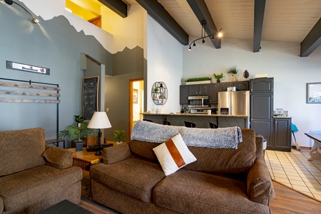 Living room interior with a brown sofa, cushion, and throw blanket. Kitchen area visible in the background with grey cabinets and stainless steel appliances. Decor includes plants and wall art.