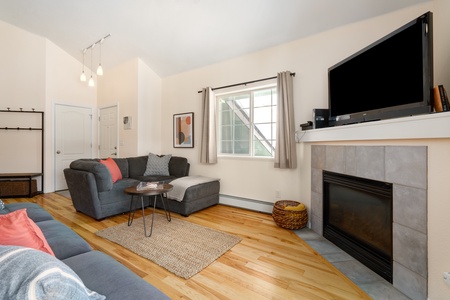 A living room with light wood flooring, gray sectional sofa, wall-mounted TV above a tile-framed fireplace, and a large window with curtains. A small coffee table sits on a woven rug.