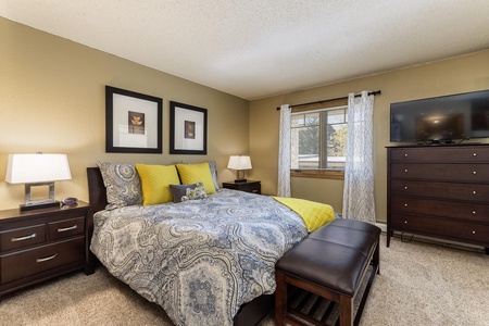 A bedroom with a double bed featuring patterned bedding and yellow accents. The room has two nightstands with lamps, a dresser with a TV, a window with white curtains, and beige carpet flooring.