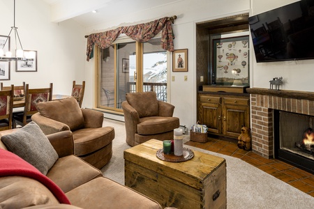 Cozy living room with a fireplace, two armchairs, a sofa, a wooden chest as a coffee table, and a TV mounted on the wall. There's a dining area with a table and chairs, and sliding doors leading outside.