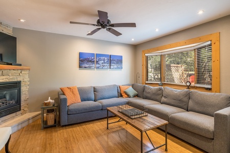 A modern living room with a gray sectional sofa, wooden coffee table, a ceiling fan, a stone fireplace, and large windows with blinds. Two landscape pictures hang above the sofa.