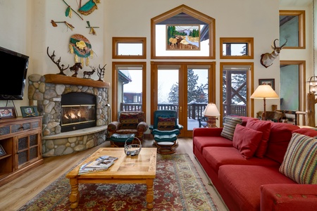 A cozy living room with a stone fireplace, red sofa, rustic furniture, and large windows showcasing a snowy landscape outside. Decor includes antler mounts and nature-themed artwork.