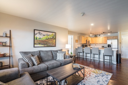 A modern, open-concept living and kitchen area features a gray sofa, wooden coffee table, colorful rug, wall art, and a kitchen with light wood cabinets and a breakfast bar with barstools.