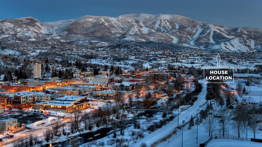 A snowy mountain town at dusk, with buildings illuminated. The location of a house is marked on the right side. Snow-covered hills and trees are visible in the background.