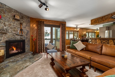 A cozy living room with a stone fireplace, brown sectional sofa, wooden coffee table, and entrance to a snow-covered outdoor area. Kitchen with bar stools is visible in the background.