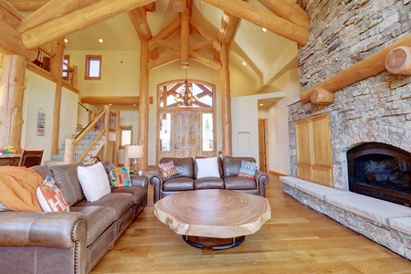 A spacious living room with wooden beams, a large stone fireplace, leather sofas, a circular wood coffee table, and a staircase leading to an upper level.