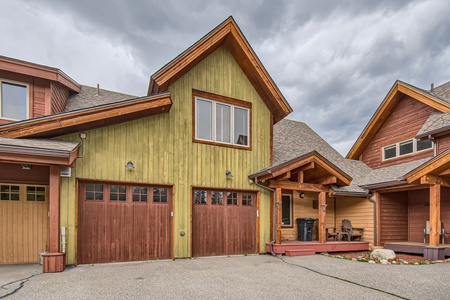 A two-story house with green and brown wooden exterior, featuring two garage doors, a front porch, and cloudy sky above.