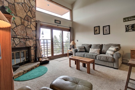 A cozy living room with a stone fireplace, gray sofa, wooden coffee table, and large windows opening to a balcony with scenic forest views.