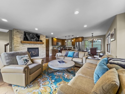 A modern living room with a stone fireplace, wall-mounted TV, sectional sofa, armchair, patterned rug, and a round coffee table. The room is adjacent to an open kitchen with wooden cabinets and stainless steel appliances.