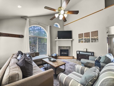 A living room with a sectional sofa and armchair arranged around a wooden coffee table, a fireplace below a TV, ceiling fan with lights, a large window with curtains, and decorative wall art.