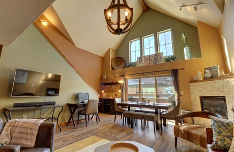 A modern living area with a high ceiling, a chandelier, a mounted TV, a dining table, a fireplace, and a staircase. The space includes wooden furniture, large windows, and various decorative items.