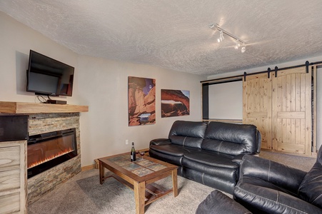 A cozy room with a mounted TV, stone fireplace, black leather couches, coffee table, and sliding wooden doors. Wall art features nature scenes. Track lighting illuminates the space.