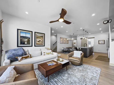 Modern living room featuring a white sofa, two armchairs, a wooden coffee table, wall art, ceiling fan, and open view of the kitchen and dining area.