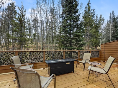 A wooden deck with four chairs surrounding a black fire table, set against a background of tall trees.