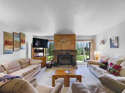 A cozy living room with light-colored couches, a coffee table, a flat-screen TV, and a stone fireplace. There is a sliding glass door leading to a garden. The walls feature a few paintings.