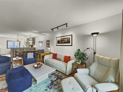 A modern living room with a sofa, armchairs, a coffee table, and wall art. A dining area and kitchen are visible in the background. The room has white walls and a mix of blue and beige furniture.