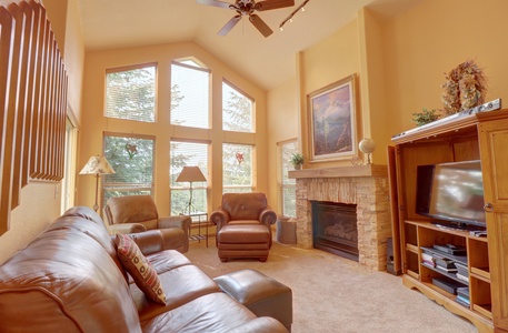 A living room with tall windows, leather furniture, a stone fireplace, a ceiling fan, a large painting, a wooden entertainment center with a TV, and decorative plants.