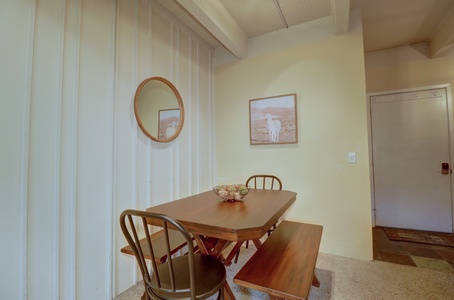 A dining area with a wooden table, three chairs, and a bench. Two framed pictures adorn the wall above the table, and a round mirror is mounted on an adjacent wall. An entry door is visible in the background.