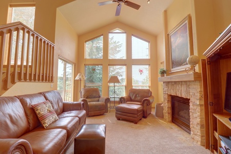 A cozy living room with high ceilings, large windows, leather furniture, a stone fireplace, and wooden accents. There is a staircase on the left and an entertainment center on the right.