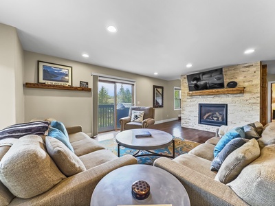 A modern living room with beige sofas, a round coffee table, a fireplace with a mounted TV above it, a wall-mounted shelf, and glass doors leading to an outdoor view.