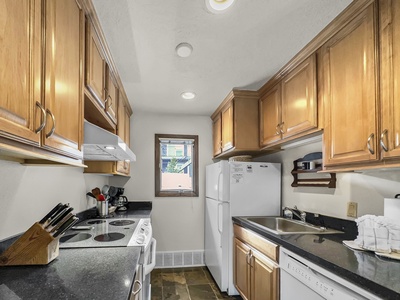 A small kitchen with wooden cabinets, black countertops, a white refrigerator, stove, and dishwasher. There's a window at the end of the room.