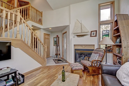 Living room with wooden flooring, a staircase, and a small fireplace. A sailboat model is mounted above the fireplace. Furniture includes a couch, TV, and a bookshelf. A bottle and glass are on the table.