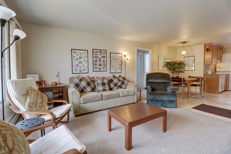 A living room features a beige sofa with plaid cushions, a green armchair, a wooden coffee table, and framed botanical prints on the wall. The adjoining dining area and kitchen are visible in the background.