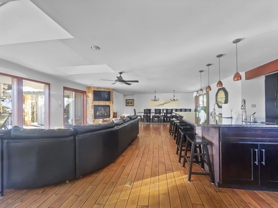 A spacious open-concept living area featuring a large black sectional, wall-mounted TV, dining table with chairs, wooden floors, and a modern kitchen with bar stools and pendant lighting.