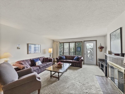 A living room with two sofas, an armchair, a coffee table, a brick fireplace, and a TV. The room has a large window and a glass door leading to an outside area.