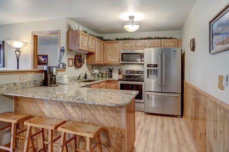 A kitchen with wooden cabinets, granite countertops, stainless steel appliances, and three wooden stools at a breakfast bar. A framed picture hangs on the wall.