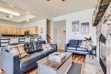 Living room with two dark leather couches, a wooden coffee table, a stone fireplace on the right, and a kitchen with wooden cabinets and a breakfast bar in the background.