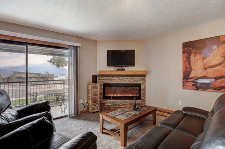 A cozy living room with a leather couch, a wall-mounted TV above a stone fireplace, a coffee table, and a painting of a canyon. Sliding glass doors open to a balcony with mountain views.