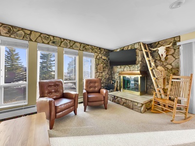 A cozy living room with stone walls, two leather armchairs, a rocking chair, a stone fireplace, and a mounted TV above. Large windows provide natural light and views of trees outside.