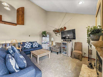 Living room with a blue couch, wooden coffee table, TV on a stand, and various decorations including plants and wall-mounted antlers. Adjacent are shelves, a chair, lamps, and a fireplace.