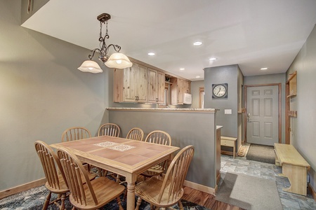 A dining area with a wooden table and six chairs. Adjacent is a kitchenette with wooden cabinets, a clock on the wall, and an entryway visible in the background.