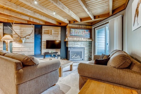 A cozy living room with wooden beamed ceiling, two sofas, a coffee table, a wall-mounted TV, a modern fireplace, deer antler decor, and sliding glass doors to a balcony or patio.