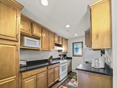 A modern kitchen with wooden cabinets, black countertops, a white stove, microwave, and refrigerator. The room has a small window and recessed lighting, along with various kitchen appliances and a colorful mat.