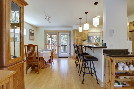 A kitchen and dining area with wooden furniture, including a high counter with bar stools, a dining table with chairs, cabinets, and a glass door leading outside.