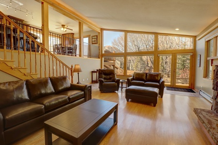 A spacious living room with wooden flooring, large windows, and leather furniture. An open staircase and dining area are visible in the background.