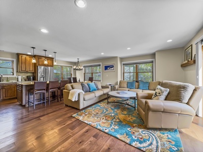 A spacious living room with a sectional sofa, colorful rug, large windows, and a connected kitchen with a breakfast bar and four stools.