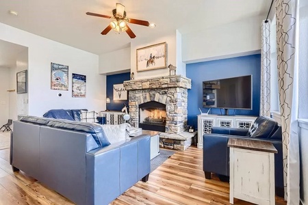 A living room with blue walls, blue sofas, a stone fireplace, a ceiling fan, a wall-mounted TV, and wooden flooring. Decor includes wall art and a framed picture above the fireplace.