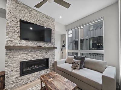 A modern living room with a stone fireplace, wall-mounted TV, gray couch, wooden coffee table, and large window with a tall building view. A ceiling fan and floor lamp are also present.