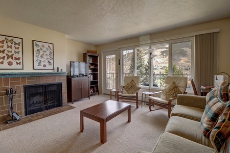 A living room with a fireplace, TV, sofa, coffee table, and two chairs. The room has large windows with a view of a hillslope outside. Butterfly-themed wall art is above the fireplace.