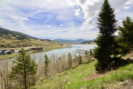 A scenic lakeside view features mountains in the background, a clear sky with scattered clouds, and pine trees on a grassy slope in the foreground.