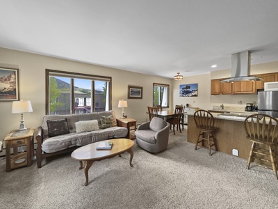 Cozy living room with a sofa, armchair, and coffee table, adjacent to dining table and kitchen with barstools. Large windows provide natural light, and framed artwork decorates the walls.